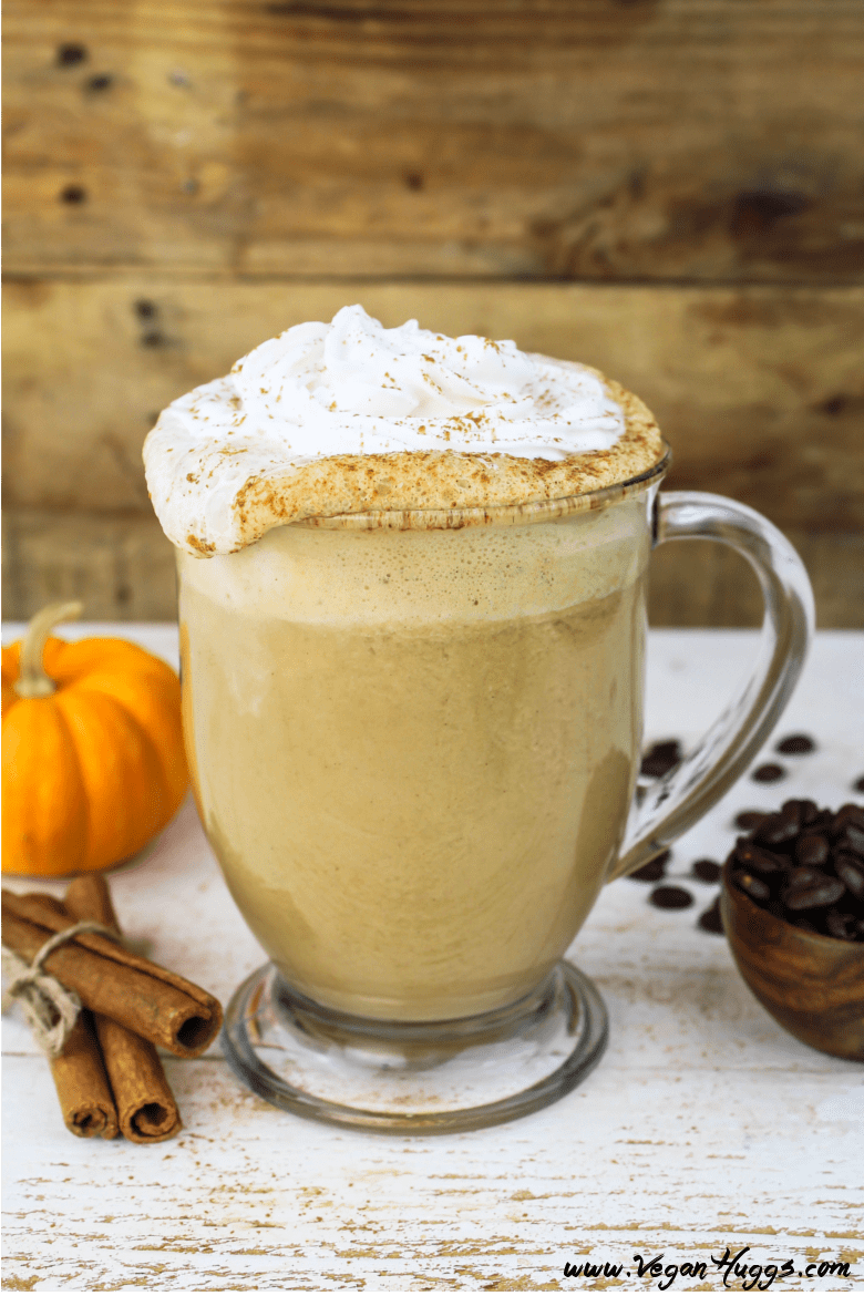 Vegan Pumpkin Spice Latte in a glass mug. Whipped cream spilling over top. Coffee beans on the side. 