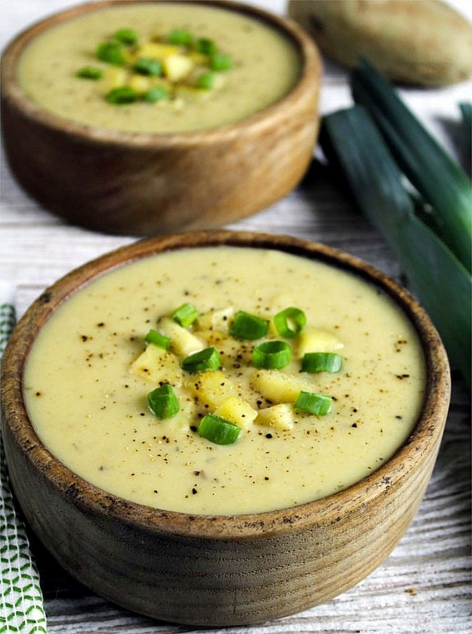 Vertical photo of two wooden bowls filled with vegan potato leek soup and topped with sliced green onion and pepper.