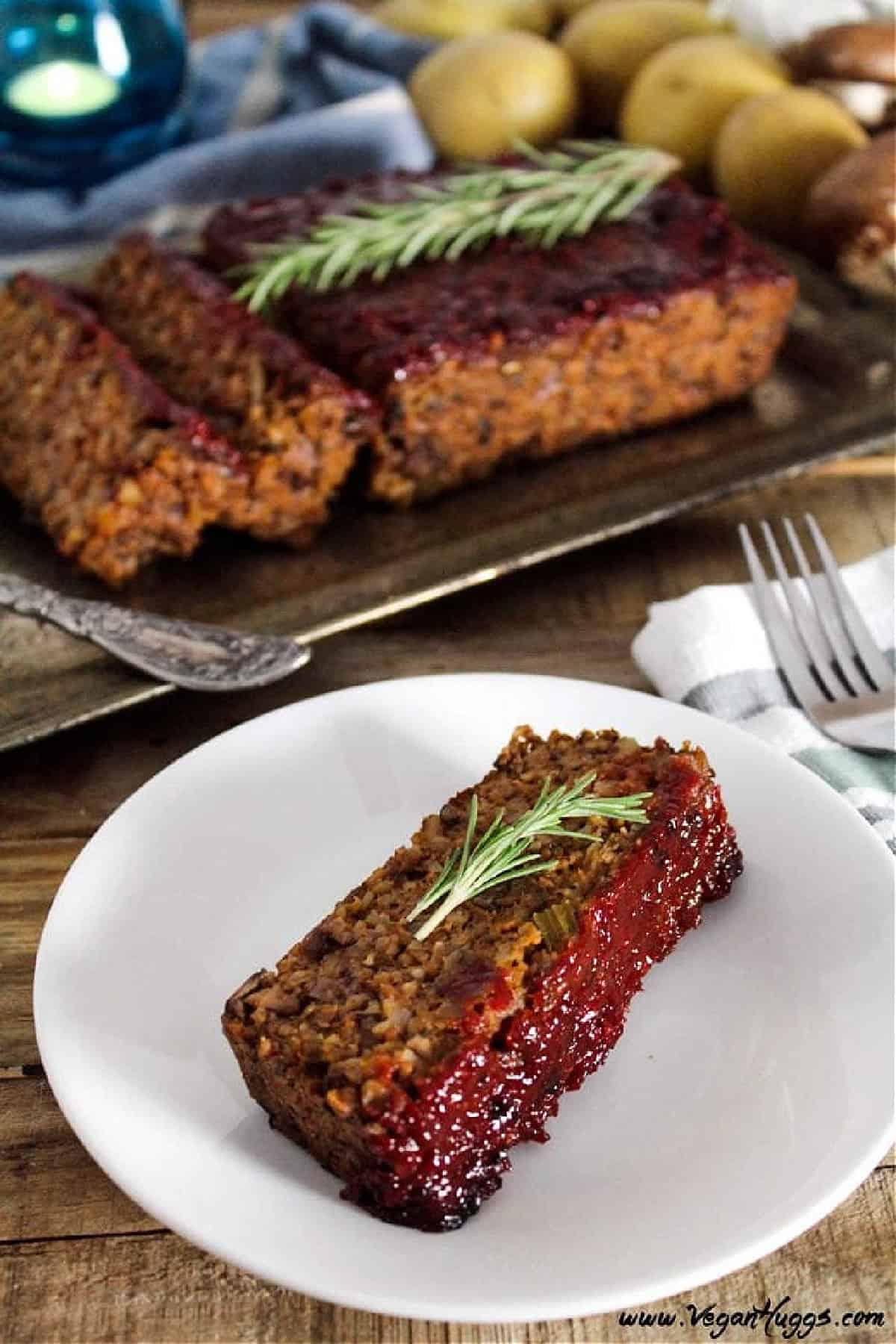 This Mushroom-Walnut Meatless Loaf w/ Ketchup Glaze is hearty, healthy & full of flavor. It will keep the vegans & non-vegans at the table together. vegan meatloaf slice on a white plate with fork on the side.