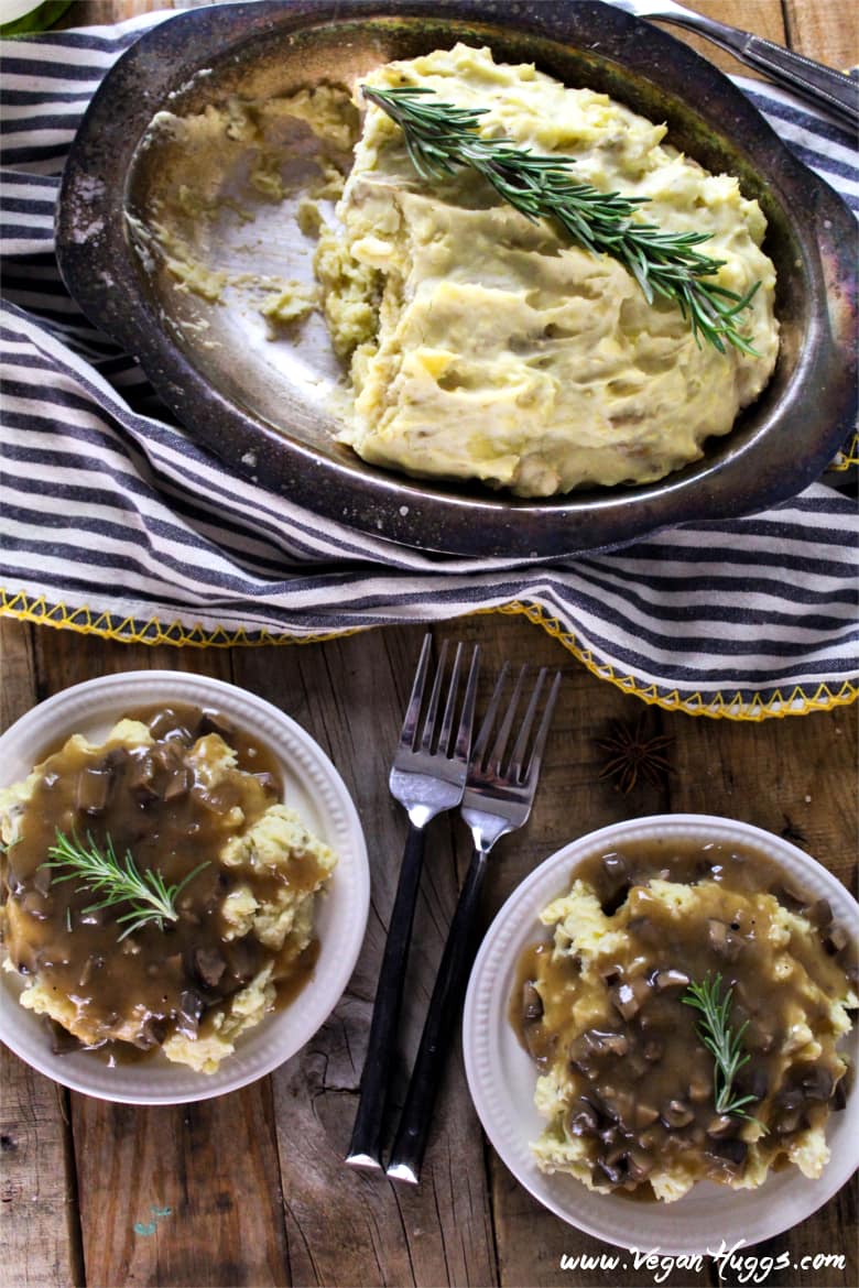 Overhead view of two plates of creamy mashed potatoes topped with mushroom gravy. Silver serving dish at the top with fresh rosemary. 