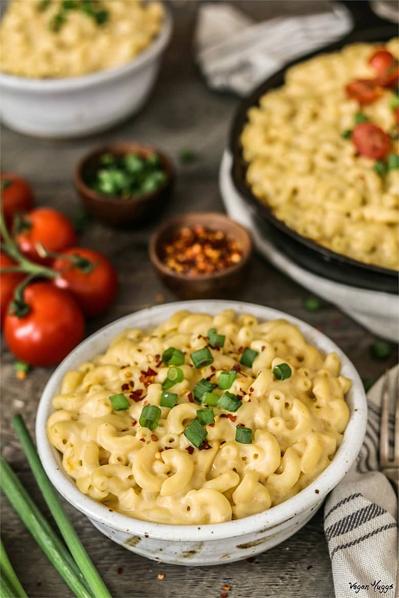 Closeup photo of Vegan Mac n Cheese in a white bowl. Filled white bowl and skillet in background. 