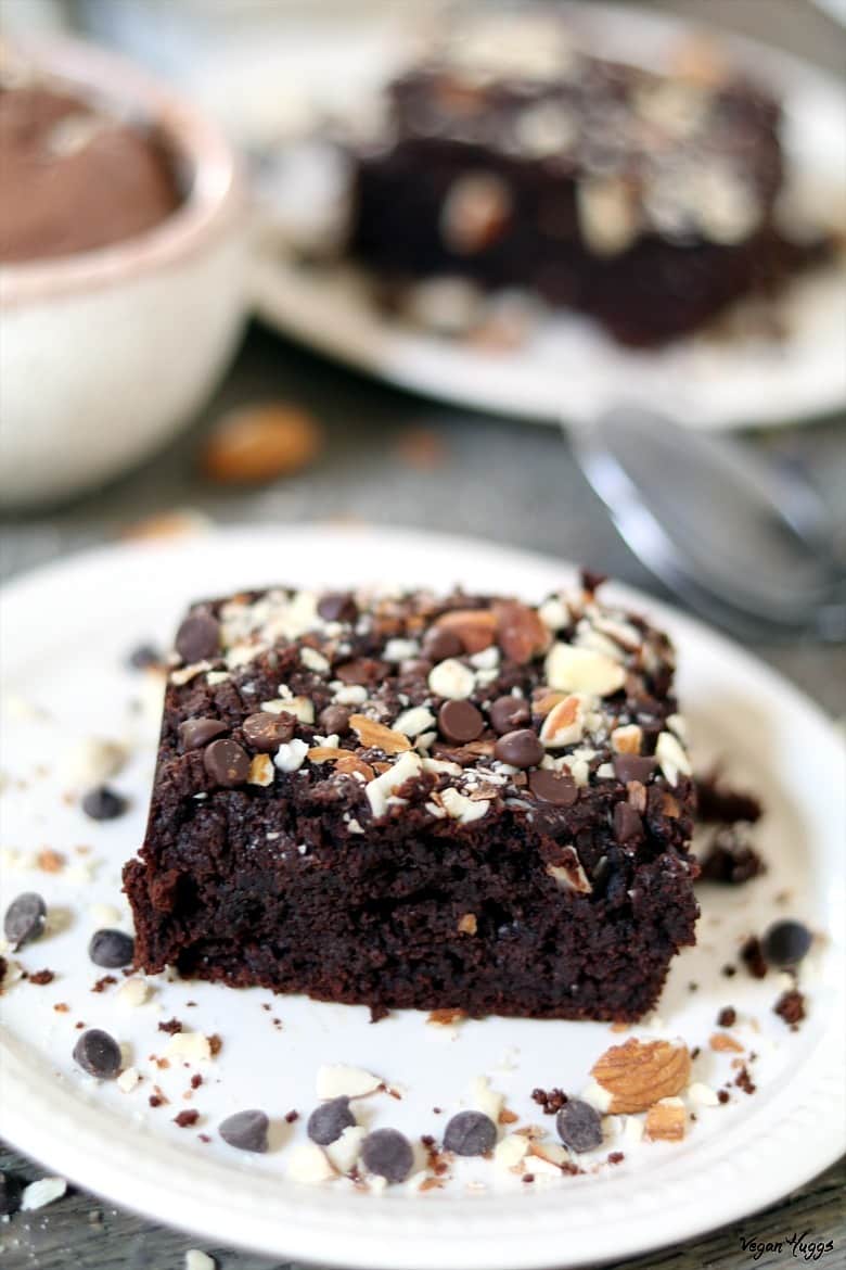 Close up view of a brownie on a white plate. Ice cream and spoon in the background. 