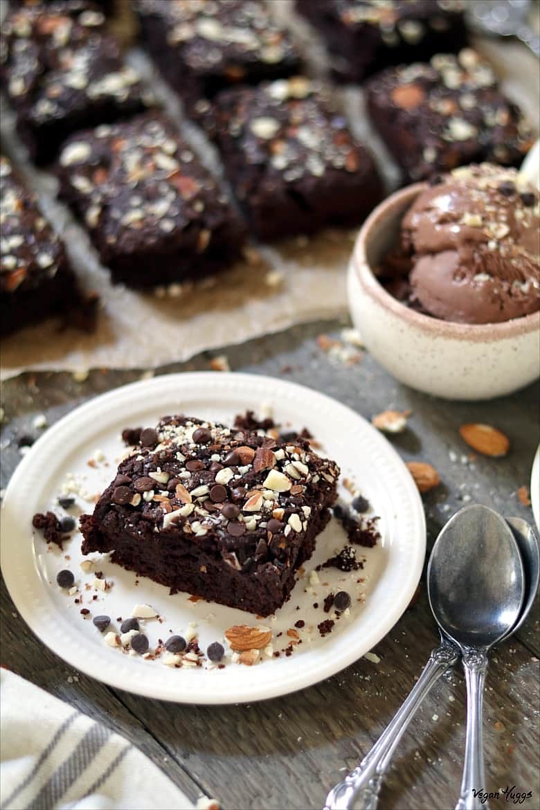 side view of a brownie on a plate with spoons in the side. Ice cream and a batch of brownies in the background.