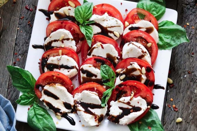 close up view of sliced vegan mozzarella cheese on a white serving plate. Topped with fresh basil, pine nuts and balsamic glaze. 