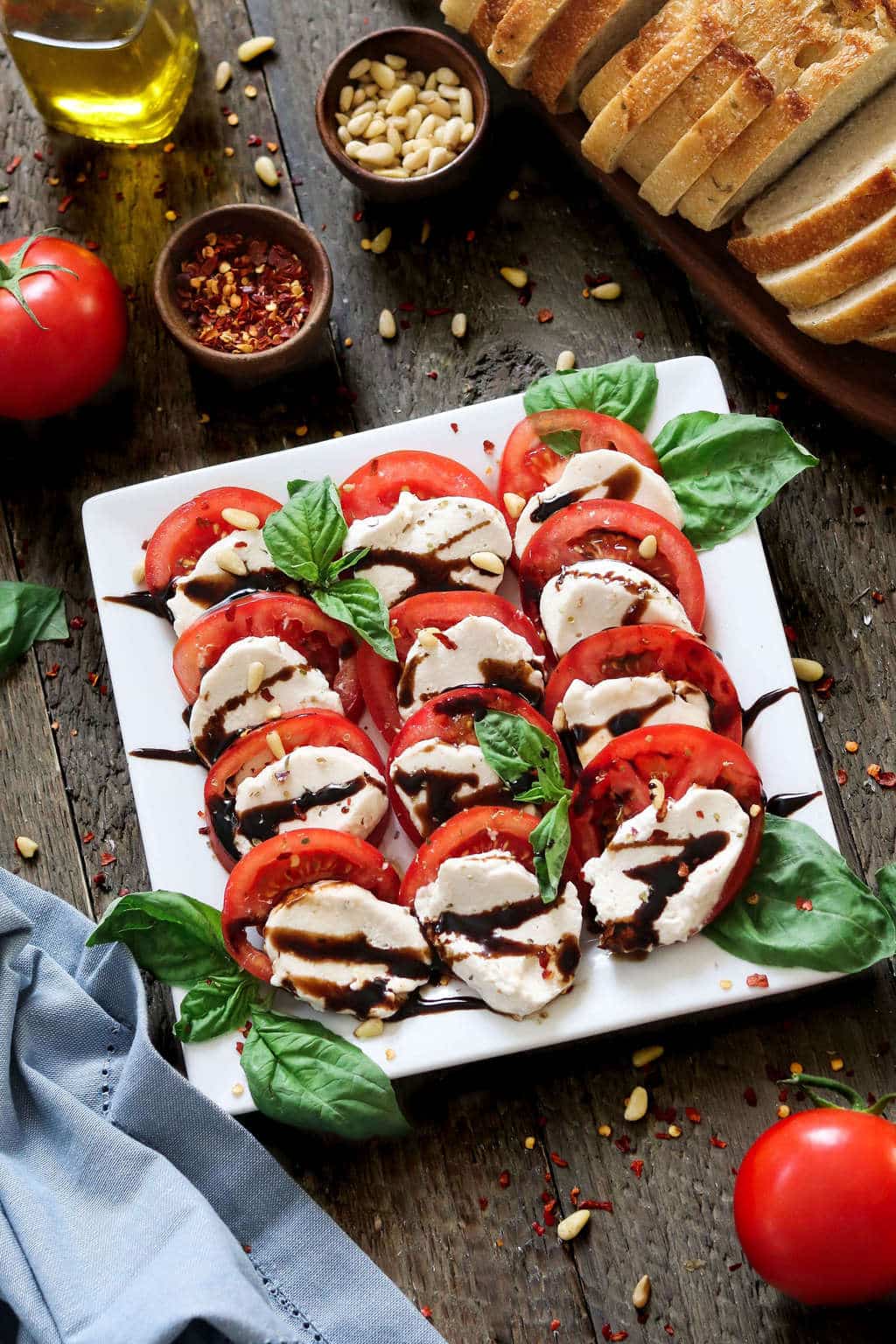 Overhead view of Vegan Mozzarella Cheese on a white serving plate. Sliced bread, tomatoes and pine nuts on the side. 