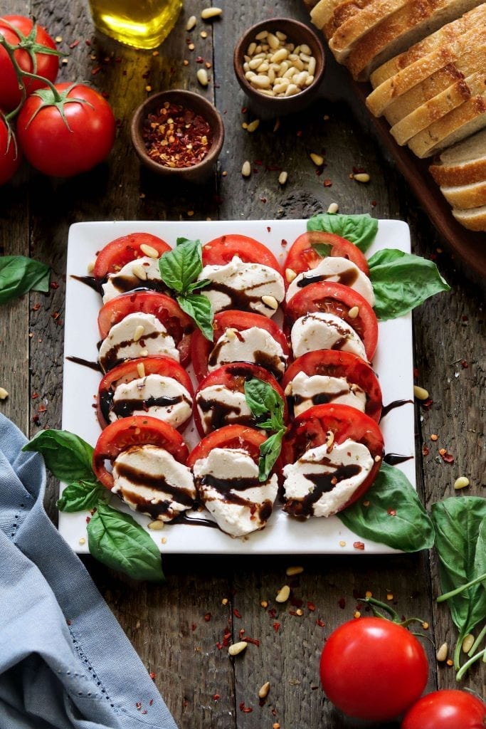 sliced vegan mozzarella cheese on top of sliced tomatoes and on a white square plate. 