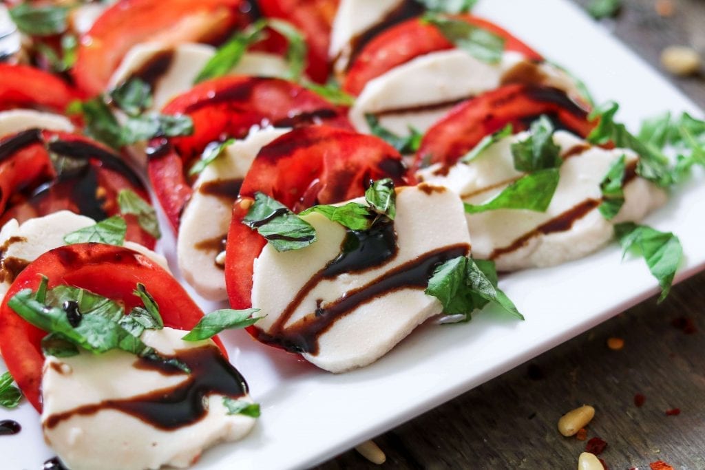 Vegan Mozzarella Cheese sliced on a white serving plate on an angled view. Topped with fresh basil and balsamic glaze. 