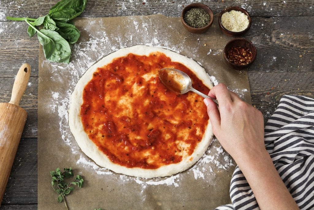 Process photo of spreading sauce on prepared pizza dough. 