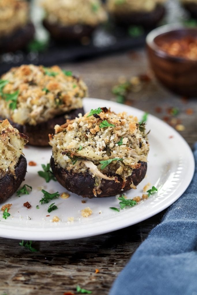 Vertical view of 3 crabless vegan stuffed mushrooms on a white plate. Topped with fresh parsley. 