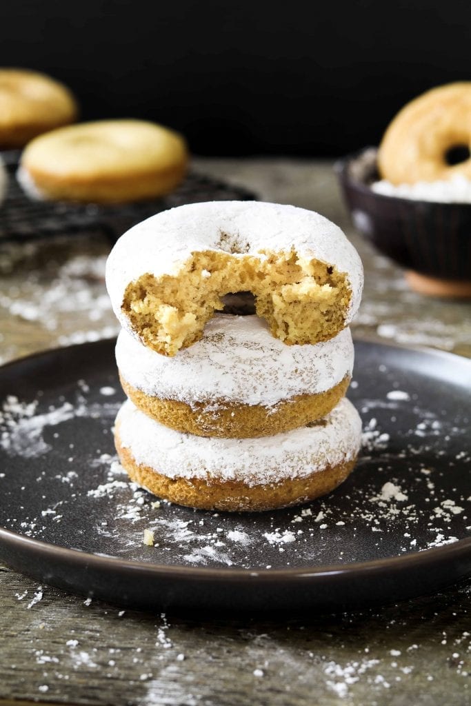 vertical photo of 3 vegan powdered donuts on a grey plate. 3 plain donuts in the back.