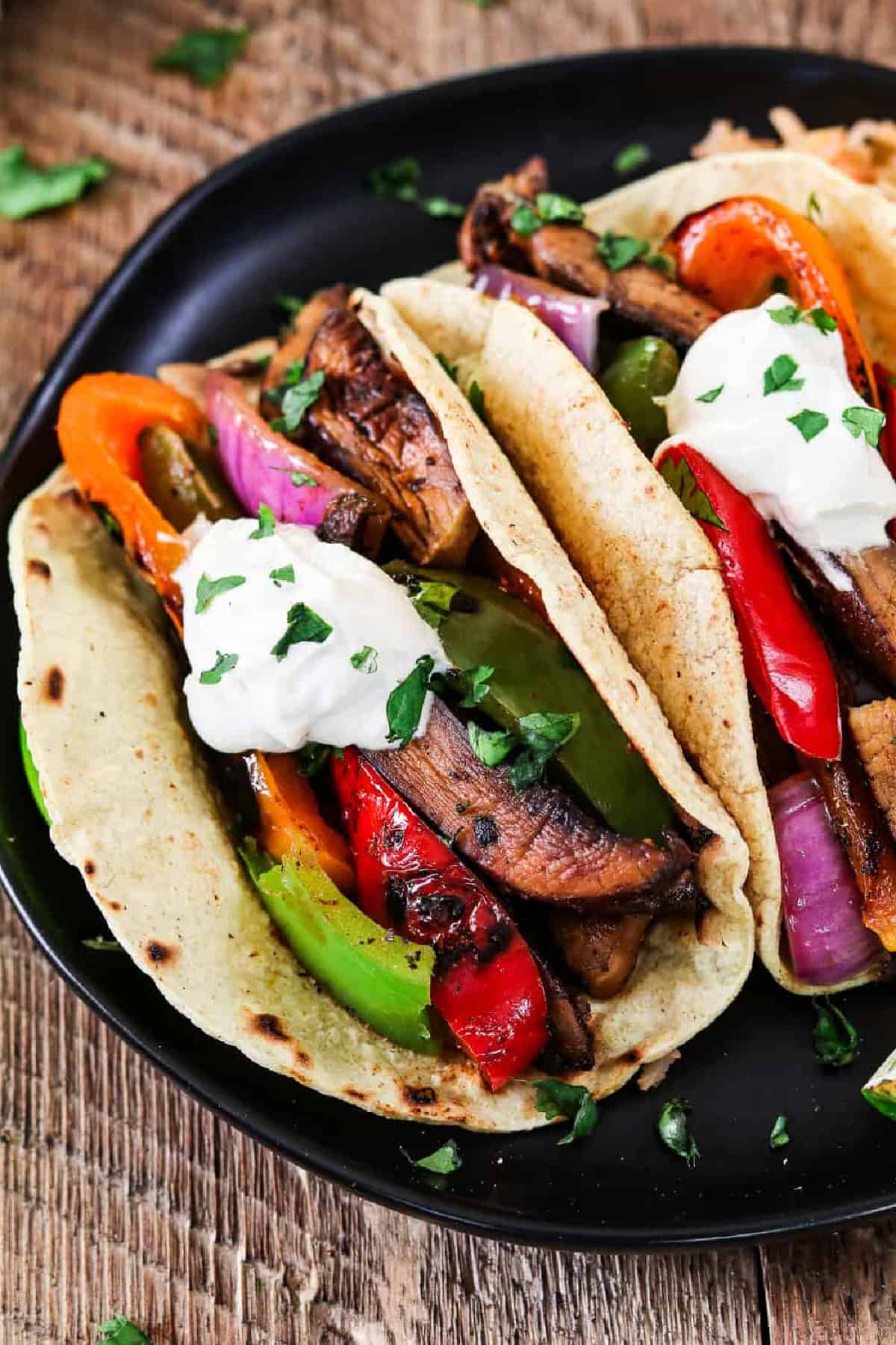 Get your weeknight sizzling In just 30 minutes with these Portobello Vegan Fajitas. They are spicy, smoky, subtly sweet and robust! The flavors come together when the seared veggies meet the homemade fajita sauce. It's mouth-watering good! closeup shot of 2 vegan fajitas, topped with vegan sour cream.