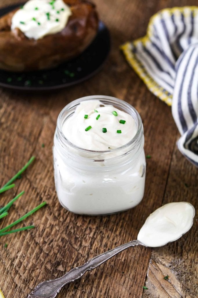 Small jar of vegan sour cream topped with chives. Spoonful of sour cream on the table and baked potato in the background. 