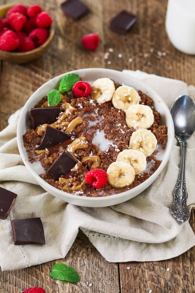 Breakfast quinoa bowl topped with bananas, chocolate and peanut butter. Raspberries in a small bowl on the side. 