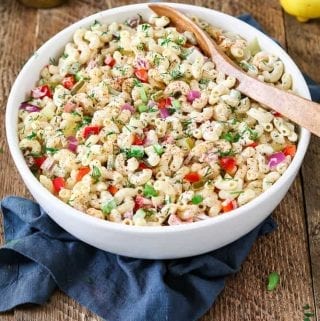 Mixed and ready to serve vegan macaroni salad in a white bowl. Blue napkin underneath and lemon wedges on the side.