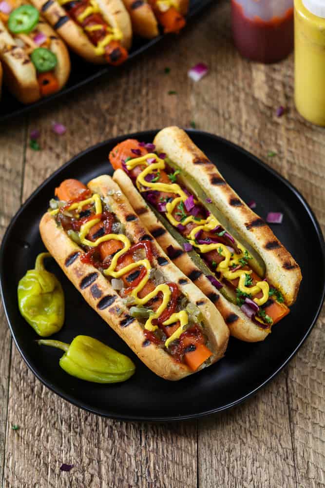 Overhead photo of two Vegan Carrot Hot Dogs on a black plate. Ketchup and mustard in the background. 