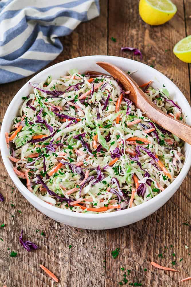 Vegan Coleslaw in a white serving bowl with a wooden spoon. Lemon in the background.