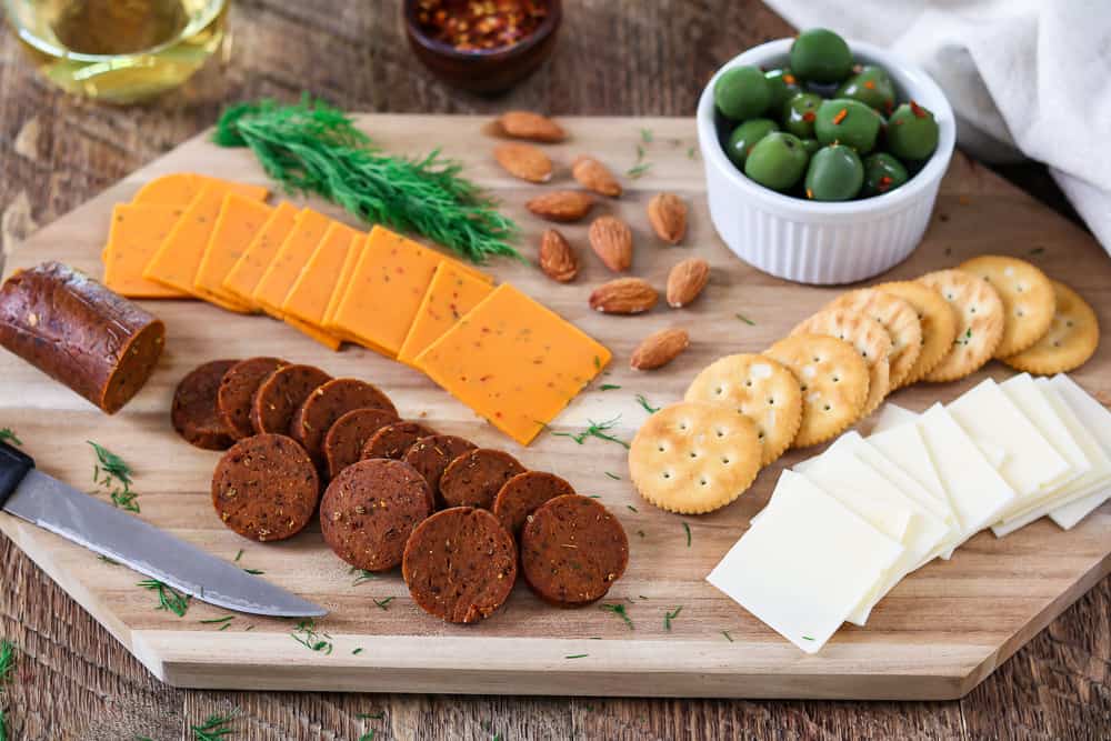 Full shot of snack board with vegan cheese, almonds, fresh dill, crackers and vegan pepperoni recipe. White wine on the side. 