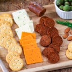 Wood cutting board with cheese, crackers, almonds and vegan pepperoni.