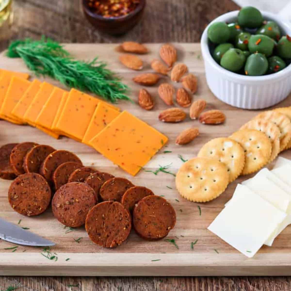 sliced vegan pepperoni on a cutting board with vegan, cheese, olives, almonds and fresh dill. 
