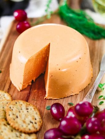 Closeup view of vegan cheddar cheese wheel. One piece cut out. Red grapes and crackers on the side.