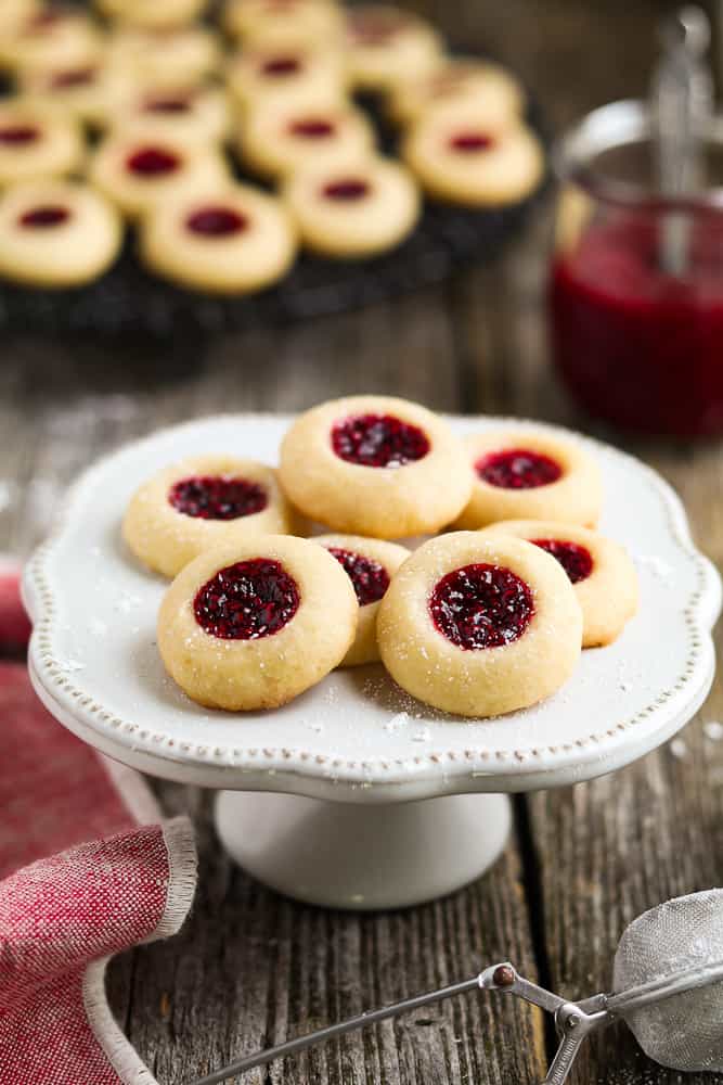 fully baked vegan thumbprint cookies on a white serving dish. Jar of jam in the background. 
