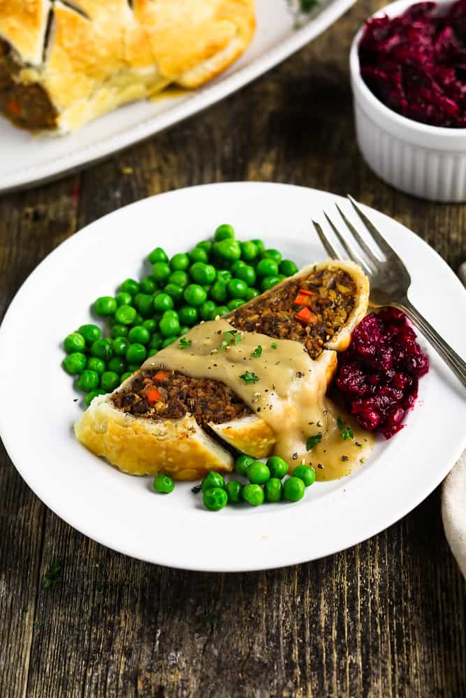 One cut piece of vegan wellington on a white plate. Peas and cranberry sauce on the side with a fork. 