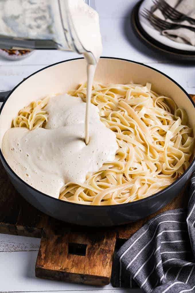 Pouring vegan alfredo sauce into a pan of cooked pasta. 