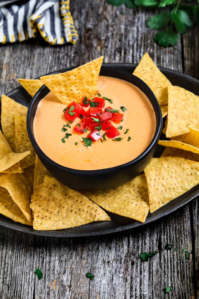 Tortilla chip sitting in a bowl of vegan nacho cheese. Striped napkin and fresh cilantro on the side.