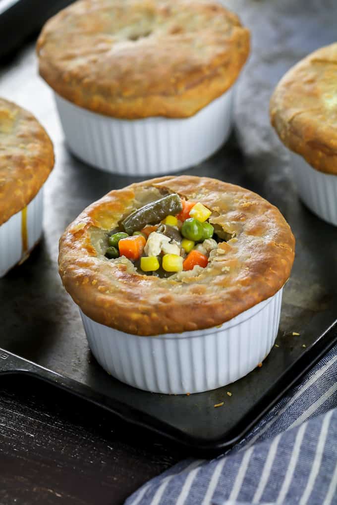 4 fully baked vegan pot pies on a baking sheet with a striped napkin on the side. One vegan pot pie is cut open on top with vegetables showing. 