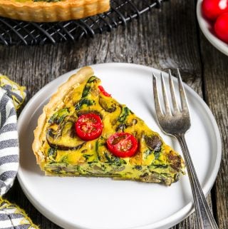 One slice of vegan quiche on a white plate. Tomatoes in a bowl on the side with full quiche.