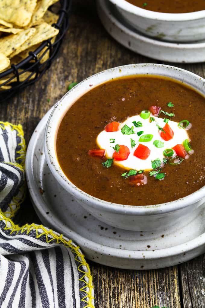 Close up view of Mexican black bean soup in a white bowl, topped with vegan sour cream, green onions and tomatoes. 