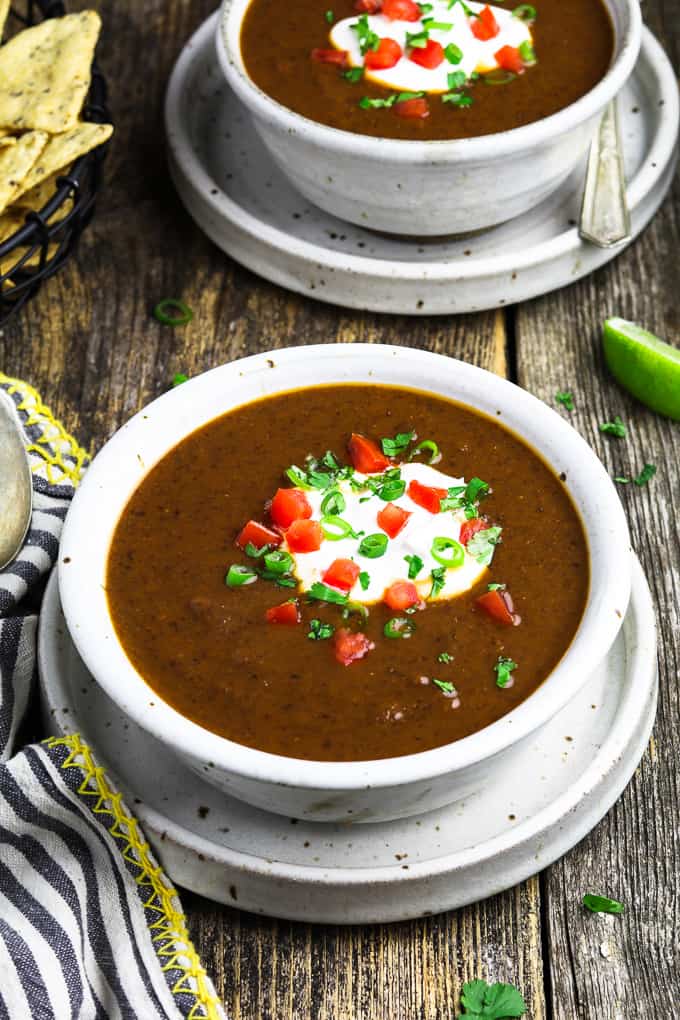 Two white bowls filled with Mexican black bean soup topped with vegan sour cream, green onion, cilantro and tomatoes. Tortilla chips on the side. 