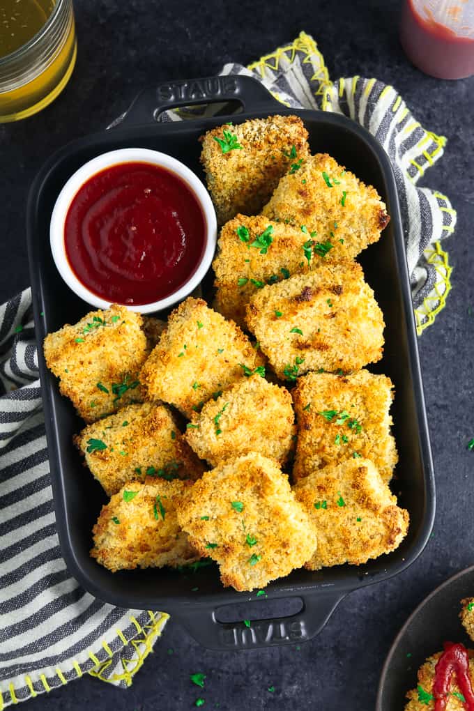 Overhead view of baked tofu nuggets in a black casserole dish with a side of ketchup.