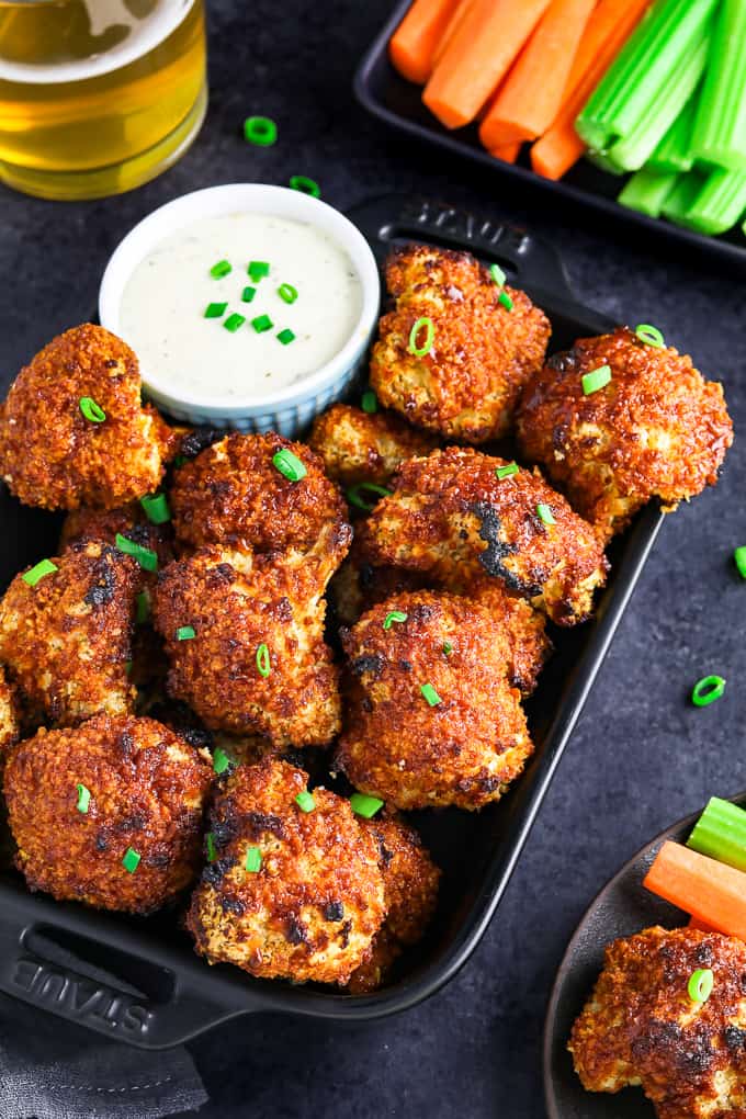 Fully baked cauliflower wings in a black casserole dish. Veggie sticks and glass of beer on the side. 