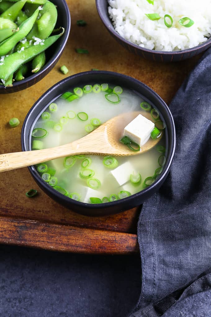 Tofu on a wooden spoon that's in a bowl of vegan miso soup. Edamame and rice on the side. 