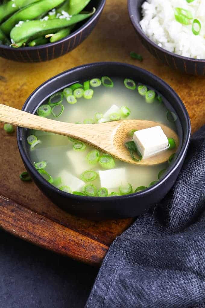 vegan miso soup in a black bowl with a wooden spoon on top. Edamame and rice on the side. 