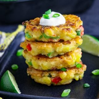 stack of four vegan corn fritters on a black plate with lime wedges on the side.