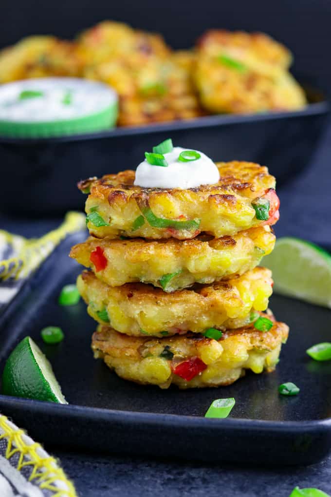 stack of four vegan corn fritters on a black plate. Topped with vegan sour cream and green onions. 