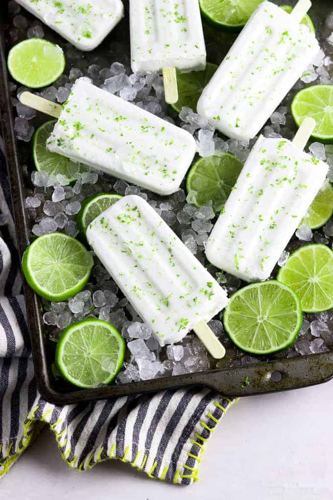 6 coconut lime popsicles on a baking tray on top of ice and limes. 