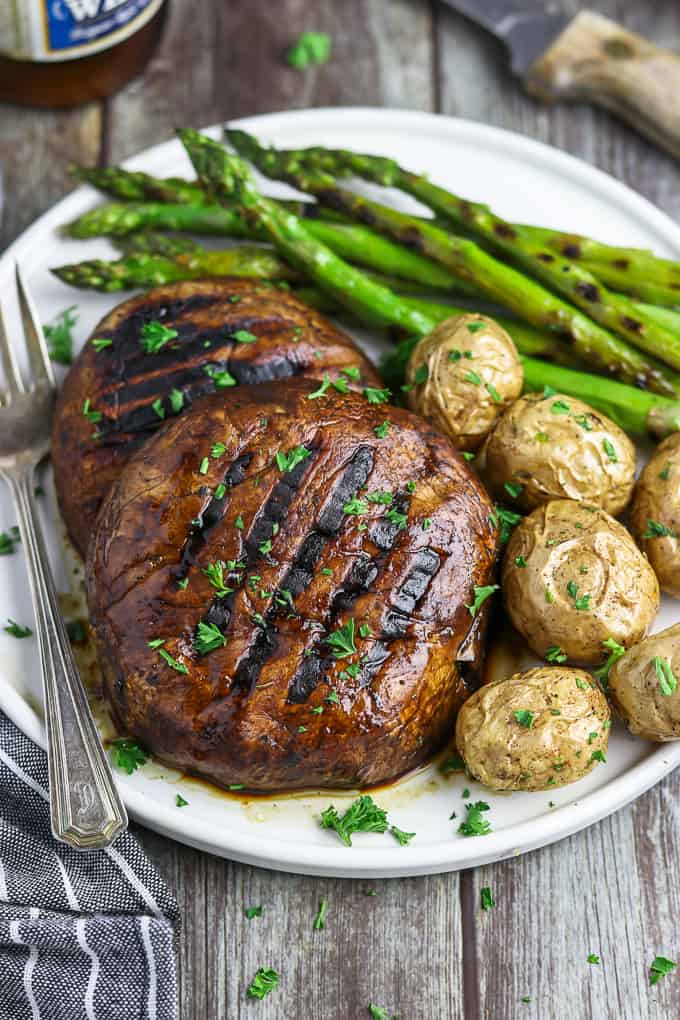 Two portobello mushroom steaks on a white plate with asparagus and potatoes on the side. 