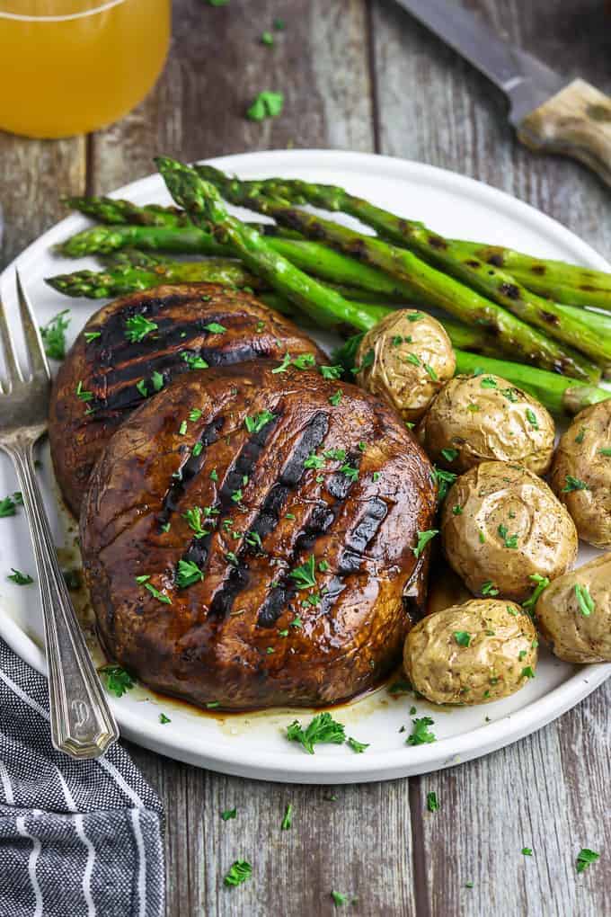 Two portobello mushrooms on a white plate with veggies and potatoes on the side. Steak knife and beverage in the background. 