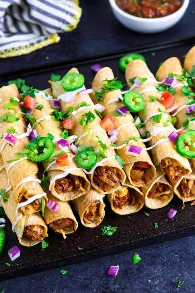 Overhead view of stacked taquitos on a black tray with salsa and napkin in the back. 