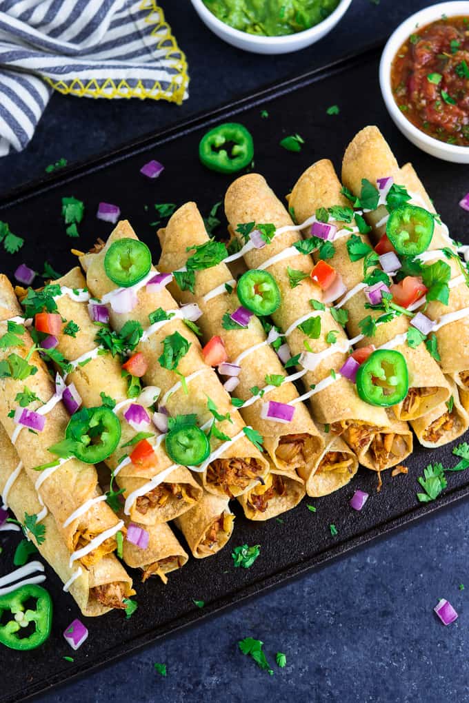 Overhead view of taquitos on a black tray with guacamole and salsa on the side. 