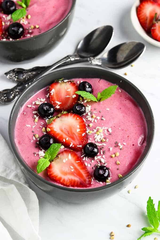 Two berry smoothie bowls on a white table with spoons in the middle. 