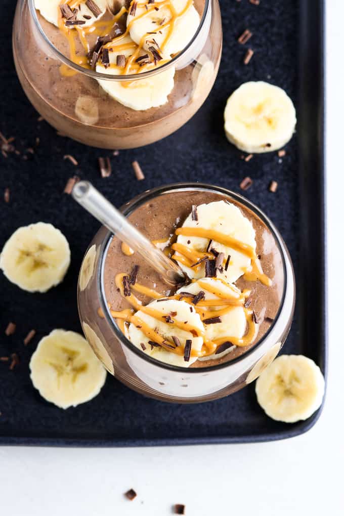 Overhead view of two smoothies on a black tray with banana slices and chocolate shavings on the side. 