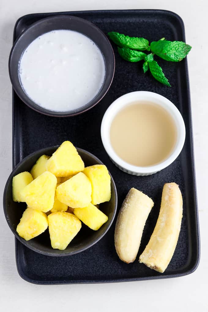 Pineapple, banana, coconut milk, fresh mint and coconut water on a black serving tray.