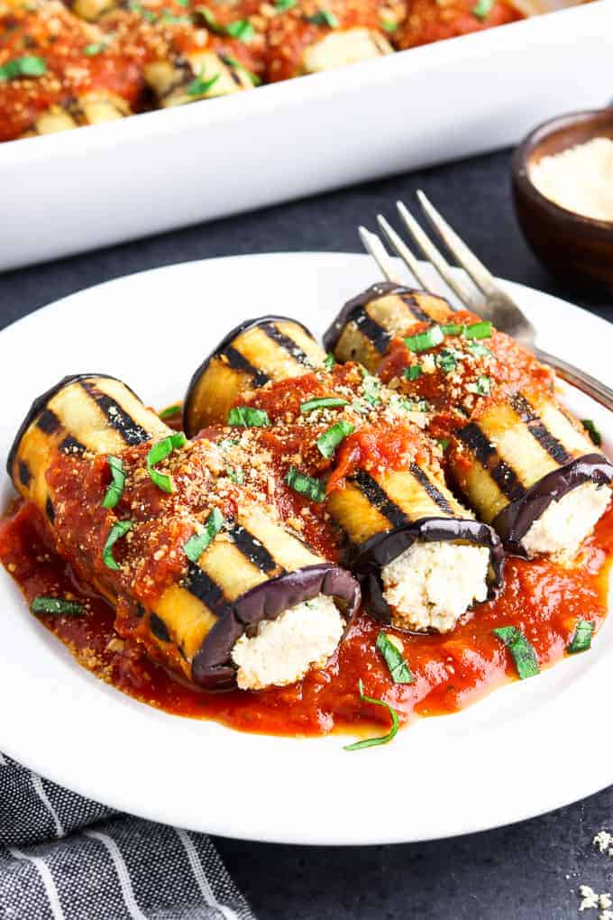 3 pieces of vegan eggplant rollatini on a white plate with fork and vegan parmesan on the side. 