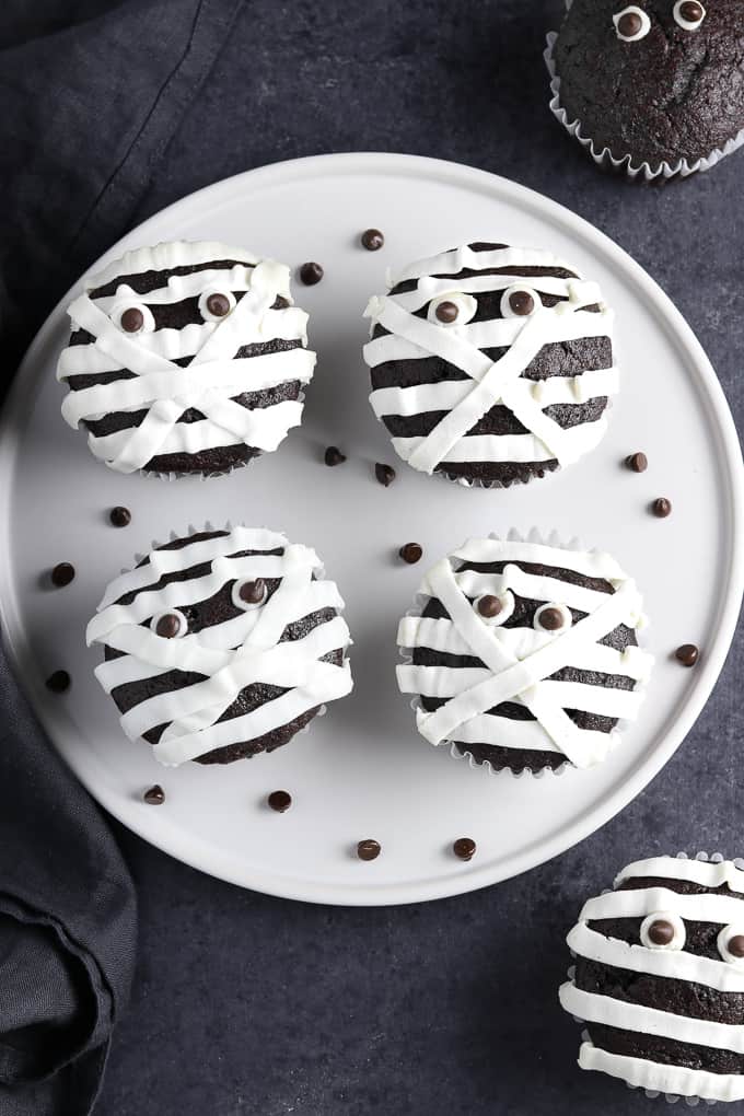 4 mummy cupcakes on a white plate with chocolate chips on the side. 