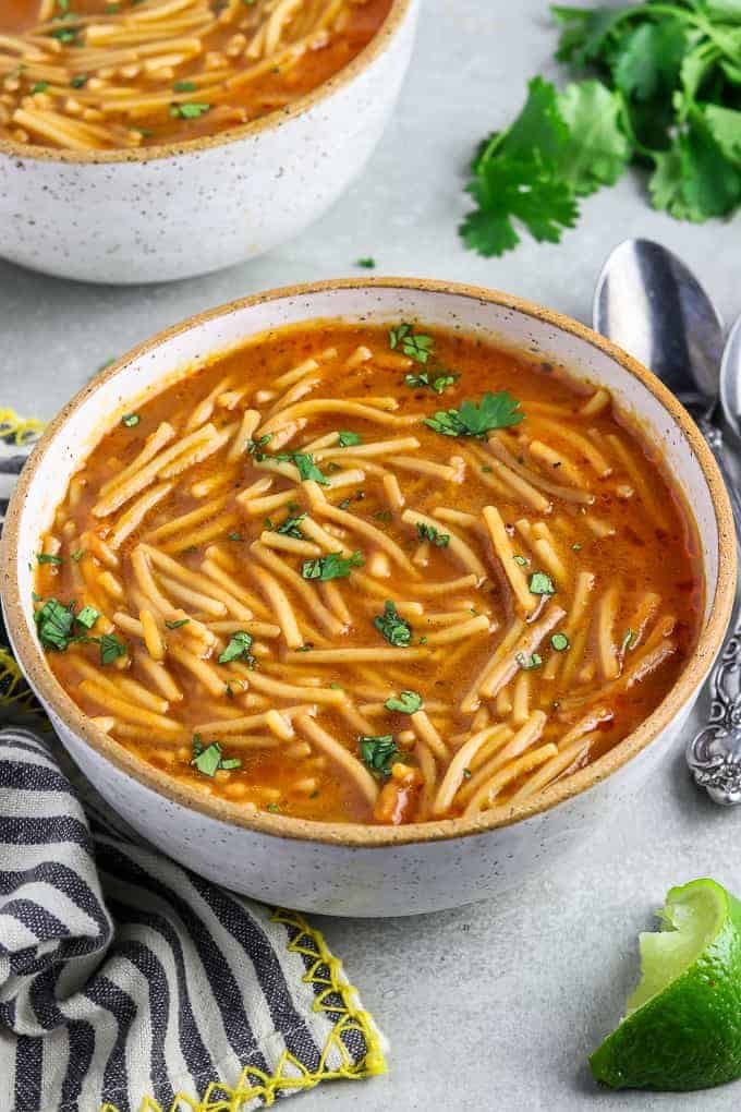 Two bowls of sopa de fideo topped with fresh cilantro. Lime, spoons and a napkin on the side. 