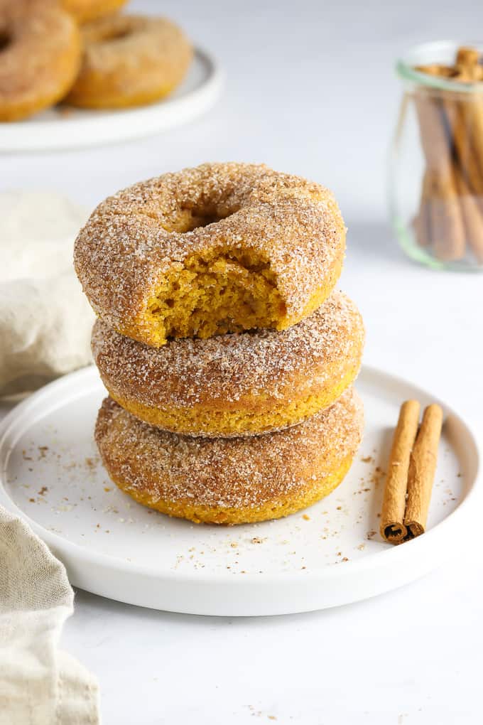 Three vegan pumpkin donuts stacked on a white plate with a cinnamon stick on the side. 